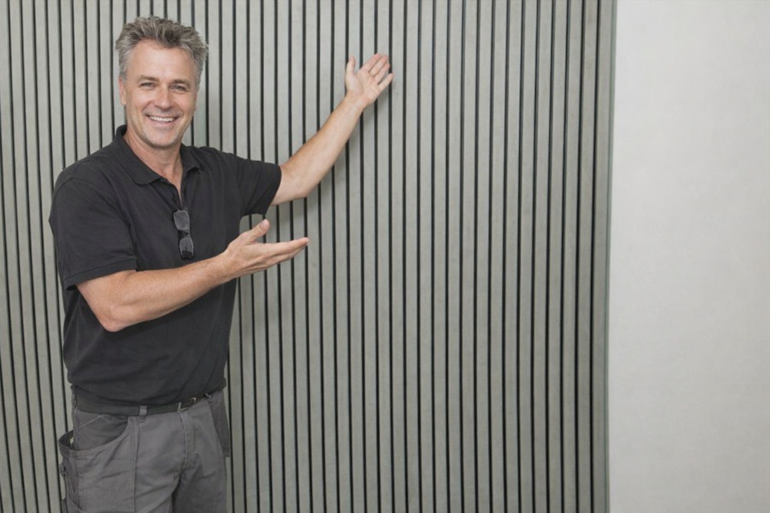 A man smiling and gesturing towards a modern grey vertical slat acoustic panel wall.